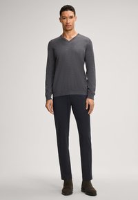 Pull gris à col en V, tissu texturé, associé à un pantalon foncé. Le mannequin porte des chaussures décontractées, les mains détendues le long du corps, sur un fond neutre.