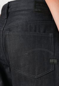 Sötét farmernadrág, hátul egy finoman ívelt öltéssel és egy "G-Star RAW" feliratú fekete bőr folttal a zsebében.