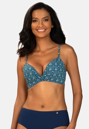 Kobieta z długimi ciemnymi włosami, uśmiechająca się, ubrana w niebieski górny strój kąpielowy w kwiatowy wzór i jednolite granatowe doły, na białym tle.