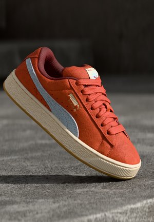 Czerwony klasyczny sneaker Puma Suede z białą podeszwą i srebrnym paskiem, umieszczony na szarej teksturowanej powierzchni.