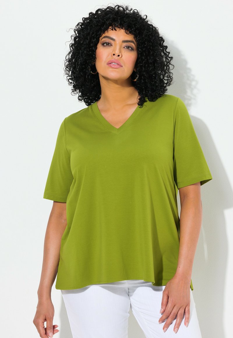 T-shirt verde oliva a maniche corte, con scollatura a V e vestibilità comoda. Tessuto morbido e leggere fessure laterali all'orlo. Pantaloni bianchi visibili.