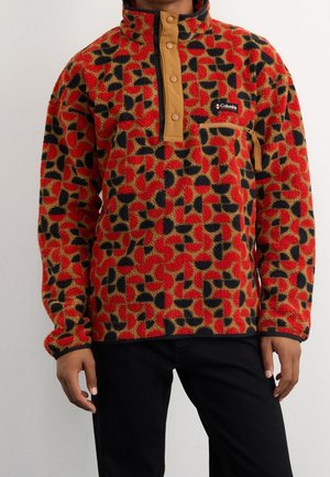 Uomo che indossa un pile con motivo rosso, nero e arancione con patta a bottoni a pressione color sabbia e tasca con cerniera, abbinato a pantaloni neri.