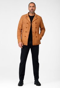 Tan læderjakke med dobbeltknapdesign, klapkrave og brune knapper. Båret over en sort skjorte med mørke jeans og støvler.