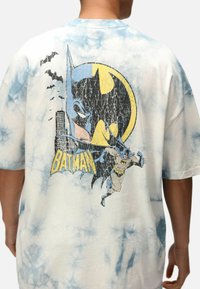 Camiseta de algodón de gran tamaño en tie-dye azul claro y blanco. Presenta un gráfico de Batman con un círculo amarillo, murciélagos negros y el texto "BATMAN" en amarillo.