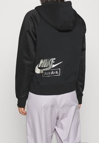 Svart huvtröja med texturerad design, huva och framträdande silverfärgad Nike-logotyp med "Just do it."-detalj på baksidan. Mjuk material.