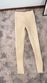 Beżowe legginsy z gładkiej, elastycznej tkaniny; dopasowany krój, wysoki stan i brak widocznych szwów, ułożone na płasko na teksturowanej jasnoszarej powierzchni.