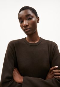 Brauner, gerippter Langarm-Sweatshirt mit Rundhalsausschnitt. Das Model hat die Arme verschränkt und zeigt die lockere Passform und die Textur des Stoffes.