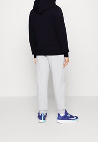 Felpa con cappuccio scura, pantaloni della tuta grigi e scarpe sportive blu con dettagli verdi, viste di spalle su uno sfondo bianco.
