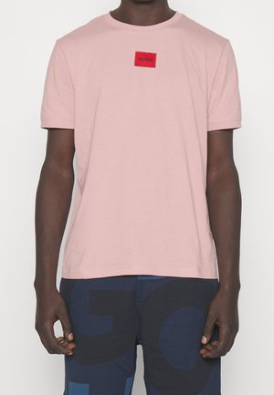 Mann trägt hellrosa kurzärmeliges T-Shirt mit rotem "HUGO"-Label auf der Brust und dunkelblaue Shorts mit großen abstrakten Mustern.
