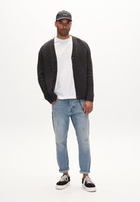 Donkergrijs gebreid vest met knopen, wit T-shirt, lichtblauwe jeans en zwarte sneakers met witte zolen, tegen een effen achtergrond.