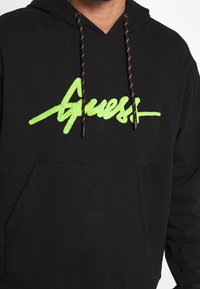 Sweat à capuche noir avec logo "Guess" texturé en vert néon sur la poitrine, doté de cordons noirs avec des rayures orange, et une poche kangourou à l'avant.