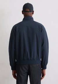 Lekka kurtka w granatowym kolorze z wysokim kołnierzem oraz elastycznym dołem i mankietami. Gładka tekstura o czystym, minimalistycznym designie.