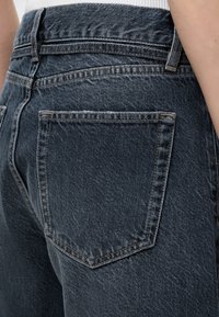 Gros plan sur un jean en denim bleu foncé montrant la poche arrière, les passants de ceinture et une partie d'un haut blanc rentré à la taille.
