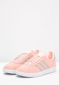 Světle růžové semišové tenisky s třemi šedými pruhy na straně, s bílou gumovou podrážkou a polstrovaným jazykem pro pohodlí.