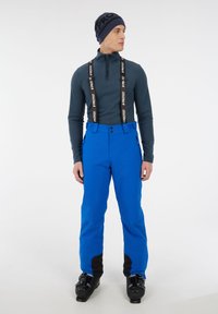 Modré lyžiarske nohavice s nastaviteľnými čiernymi kšandami, dvoma bočnými vreckami a elegantným dizajnom. Skombinované s tmavým dlhým rukávom a čiernymi lyžiarskymi topánkami.