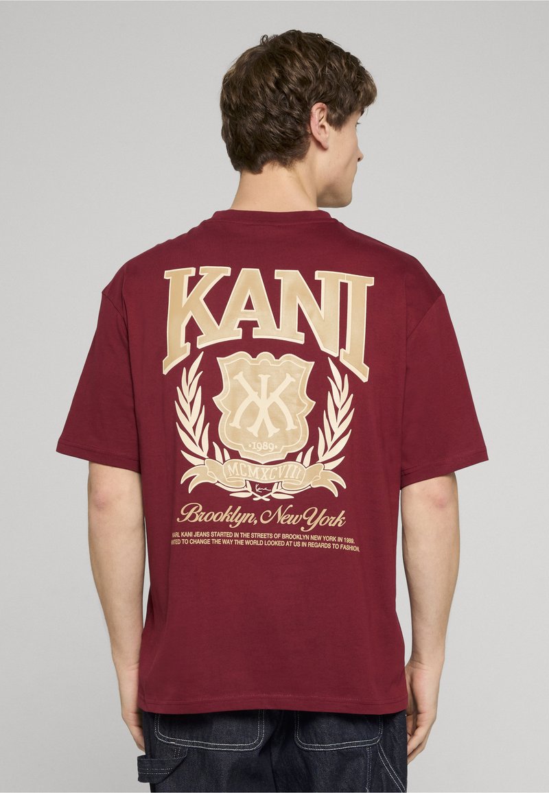 Brūna kokvilnas T-krekle ar lielu krēmkrāsas grafiku uz muguras, kas satur tekstu un emblēmu. Īsas piedurknes un brīvs piegriezums.