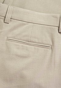 Pantaloni in tessuto beige con davanti piatto, passanti per cintura e una tasca posteriore a filetto singolo con un dettaglio di cucitura sottile. Texture liscia.