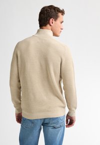 Beige stickad tröja med hög krage och ribbad textur, i kombination med blå jeans. Tröjan har långa ärmar och en figurnära design.