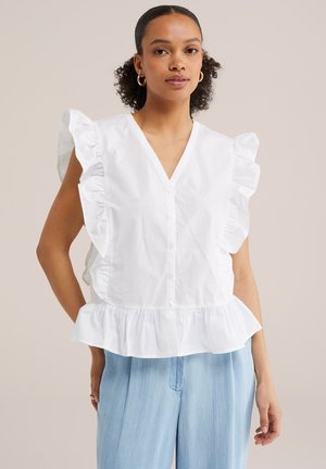 Žena s kučeravými vlasmi, ktorá nosí zlaté kruhové náušnice, bielu volánikovú tielkovú blúzku s gombíkmi a svetlomodré nohavice s vysokým pásom.