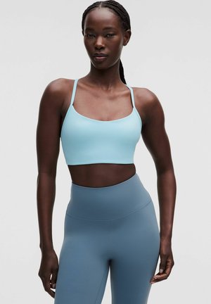 Fit kvinde med mørk hud iført en lyseblå sports-bh og højtaljede blå leggings, står mod en ensfarvet baggrund.