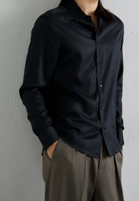 Camicia nera con bottoni realizzata in tessuto morbido, caratterizzata da un colletto a punta e maniche lunghe, abbinata a pantaloni a vita alta di colore marrone chiaro.