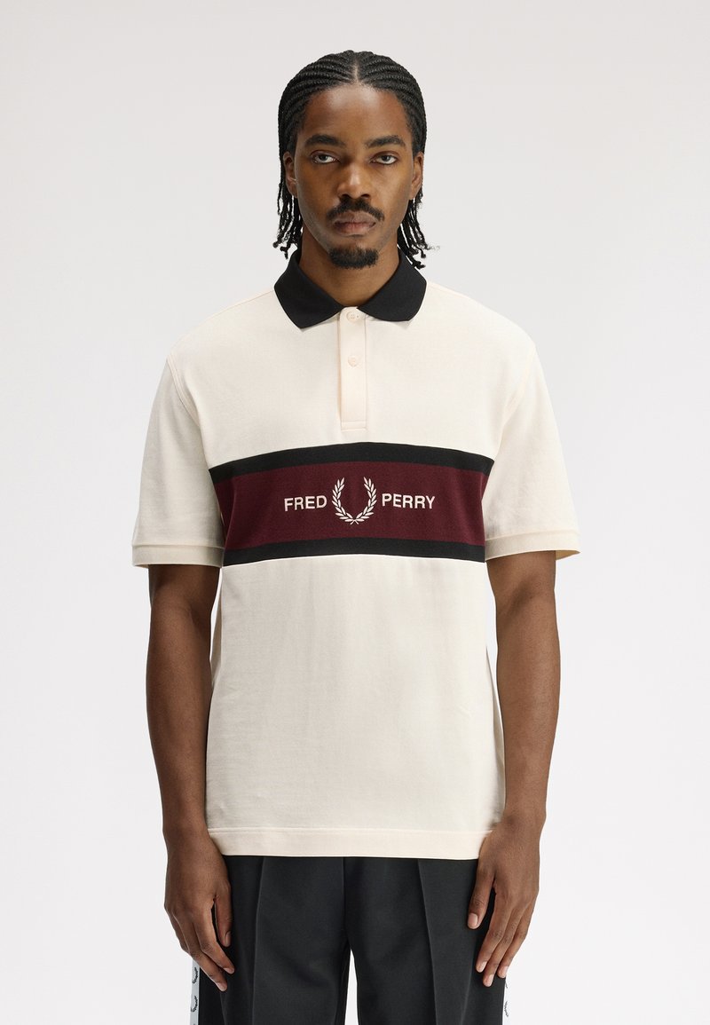 Homme aux cheveux tressés portant un polo crème avec col noir et bande bordeaux portant l'inscription "Fred Perry", debout devant un fond uni.