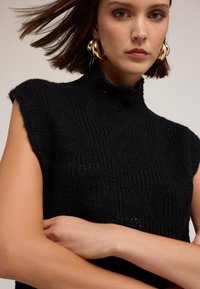 Svart stickad ärmlös topp med hög krage, med texturerade mönster och lätt genombrutet stickmönster. Guldiga hoop-örhängen ger en accent.