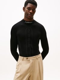 Chemise noire ajustée à manches longues avec des détails de logo sur le col, associée à un pantalon large beige clair, créant un look contrasté.