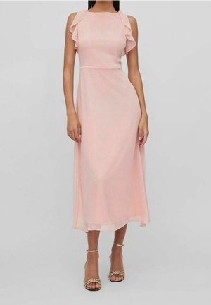 Femme portant une robe rose sans manches, longue jusqu'aux chevilles, avec des détails à volants sur les épaules et des talons ouverts dorés, debout devant un fond uni.