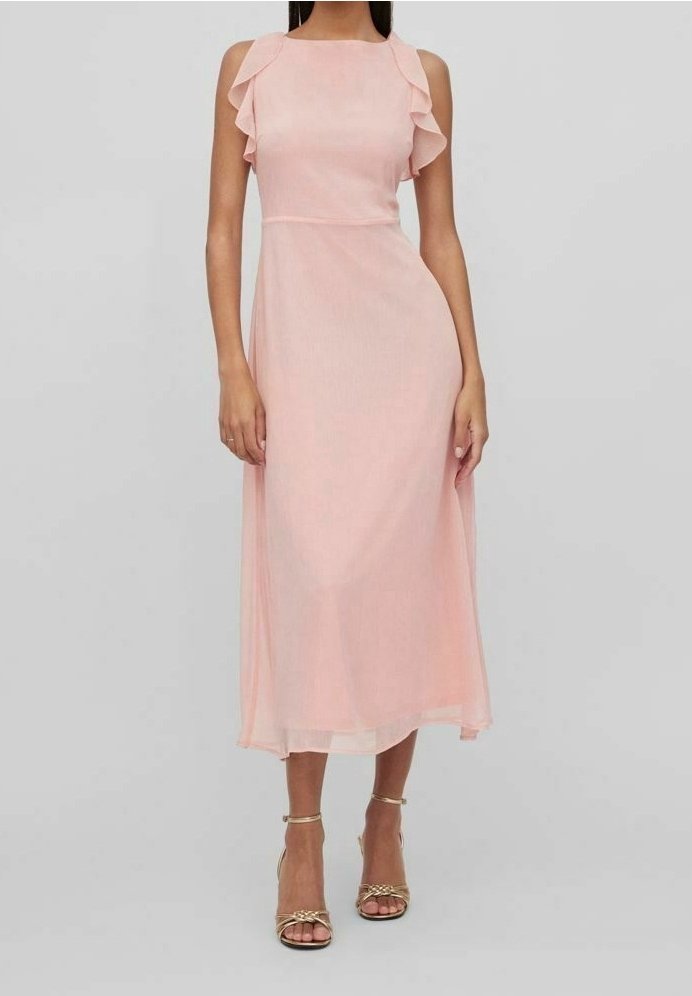 Femme portant une robe rose sans manches, longue jusqu'aux chevilles, avec des détails à volants sur les épaules et des talons ouverts dorés, debout devant un fond uni.