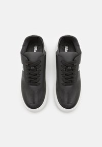Zapatillas negras con una textura suave, lados perforados y diseño de cordones. Las suelas blancas contrastan con la parte superior oscura, que presenta una etiqueta de marca.