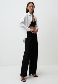 Pantalons noirs taille haute associés à un top noir court et une chemise blanche à boutonnage, présentant une coupe décontractée et un léger brillant. Talons hauts noirs à lanières.