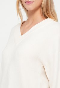 Krémový, mäkký pletený sveter s výstrihom do V a voľným strihom. Obsahuje rebrované detaily na výstrihu a jemnú textúru.