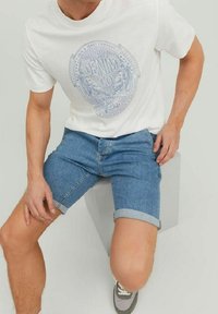 Camiseta blanca de manga corta con un diseño gráfico azul, combinada con pantalones cortos de mezclilla de color azul claro. Modelo sentado sobre una superficie gris, luciendo zapatillas grises.