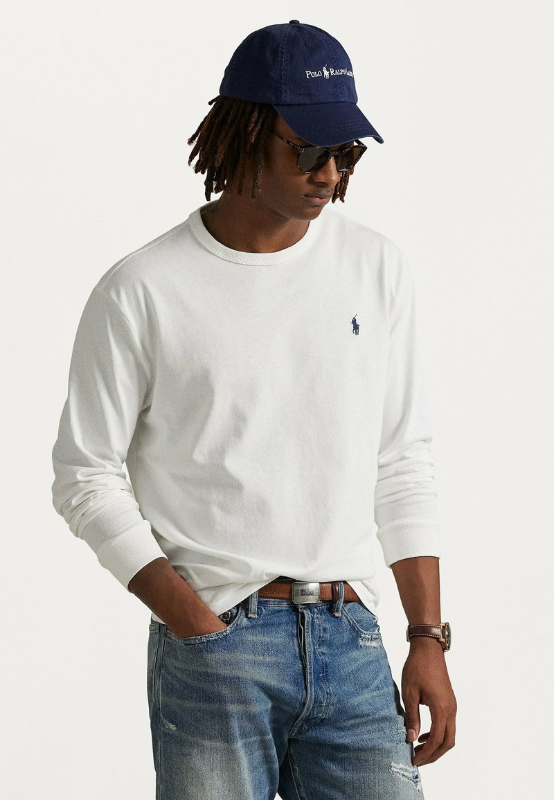 T-shirt bianco a maniche lunghe con un piccolo logo blu navy. Abbinato a jeans blu sbiaditi e un cappellino blu navy con testo bianco. Orologio da polso visibile.