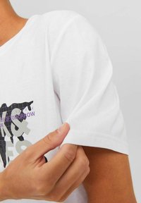 Camiseta de algodón blanca con un estampado gráfico en negro y púrpura. La manga está ligeramente enrollada, mostrando una textura suave y costuras bien definidas.