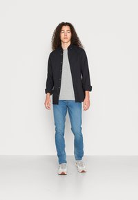 Zwart overhemd met knopen over een grijze T-shirt, gecombineerd met een blauwe spijkerbroek en grijze sportschoenen. Krullend haar, staand tegen een witte achtergrond.