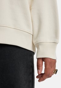 Φούτερ σε εκρού χρώμα με υφή υφάσματος και ριμπ μανσέτες, σε συνδυασμό με σκουρόχρωμο τζιν παντελόνι και δαχτυλίδι με χρυσές λεπτομέρειες στο χέρι.