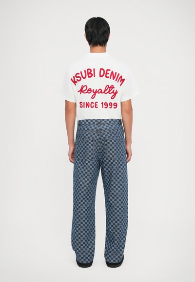 Tricou alb cu text roșu "KSUBI DENIM Royalty SINCE 1999" pe spate, asociat cu pantaloni din denim albastru cu model, cu o croială largă.