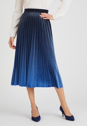 Jupe midi plissée en velours bleu dégradé, avec une taille ajustée et une texture douce. Associée à des chaussures à talons hauts bleu foncé.