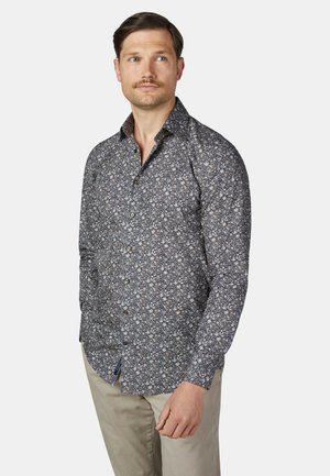 Man draagt een lange mouw donker gebloemde blouse met knopen en beige broek, kijkt iets naar links tegen een effen lichte achtergrond.