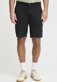 Black knee-length shorts with a smooth texture, a button closure, and belt loops. Paired with beige sneakers and white socks.