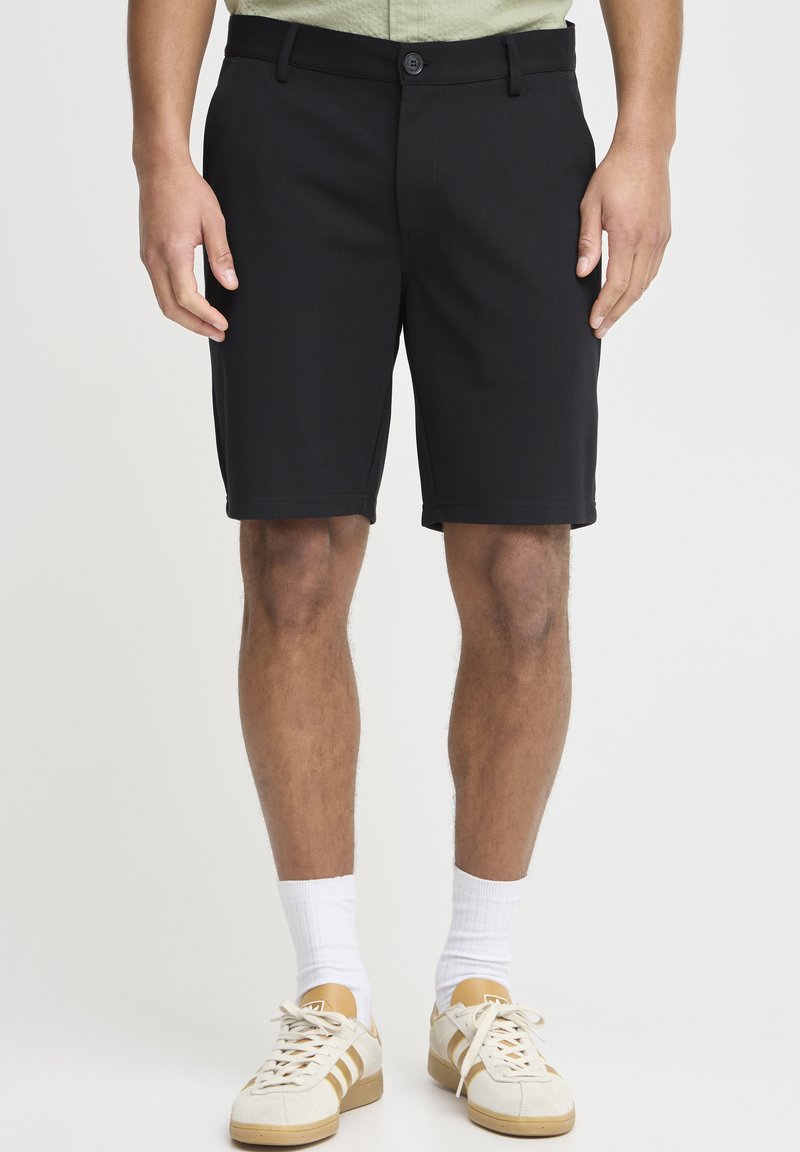 Black knee-length shorts with a smooth texture, a button closure, and belt loops. Paired with beige sneakers and white socks.