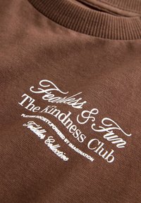 Hnědá mikina se bílým nápisem "Nebojácný a Zábavný Kluba Laskavosti" s žebrovaným límcem a měkkou strukturou.