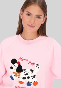 Sudadera rosa con cuello redondo que presenta un diseño de perro esponjoso en blanco y negro con acentos coloridos, texto bordado y una textura suave.