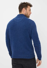 Maglione blu lavorato a maglia con collo a scialle; tessuto strutturato; polsini e orlo a coste; indossato con jeans neri, visto da dietro.