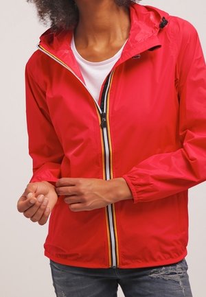 Person trägt eine rote Kapuzenjacke mit Reißverschluss und gestreiftem Reißverschluss-Detail, passt den Ärmel an, kombiniert mit abgenutzten blauen Jeans.