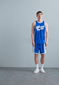 Blå sports tanktop med hvit skrift og grafikk, parret med matchende blå shorts. Begge har kontrasterende hvite detaljer. Hvite joggesko.