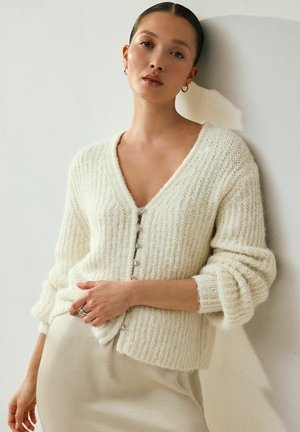 Cardigan crem cu nervuri, cu decolteu în V și nasturi argintii, fabricat dintr-un material moale și texturat. Mâneci lungi, lejere și tiv scurt.