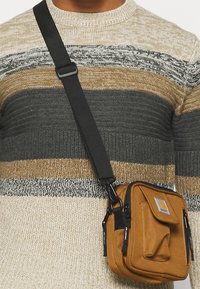 Bruine stoffen crossbodytas met meerdere ritsvakken en een zachte textuur, met een logopatch en verstelbare zwarte band.